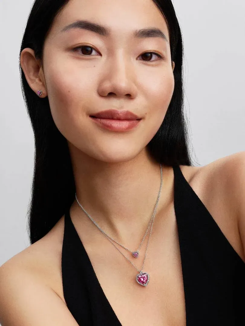 Gros plan d'un mannequin portant un collier cœur rose et des boucles d'oreilles assorties, fond neutre clair.