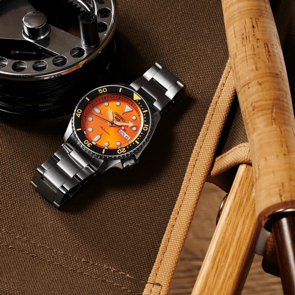 Reloj en plano cenital diagonal sobre fondo de lona marrón, junto a accesorios metálicos y de madera.