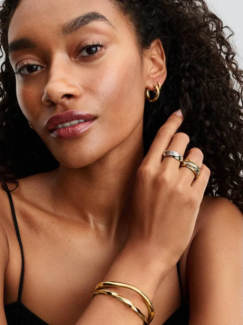 Close-up van model met gouden oorringen, ringen en een armband tegen een neutrale achtergrond.