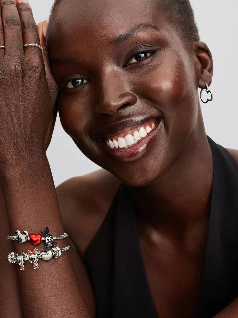 Close-up of smiling model wearing Minnie Mouse charms on wrist and silver earrings, neutral background.