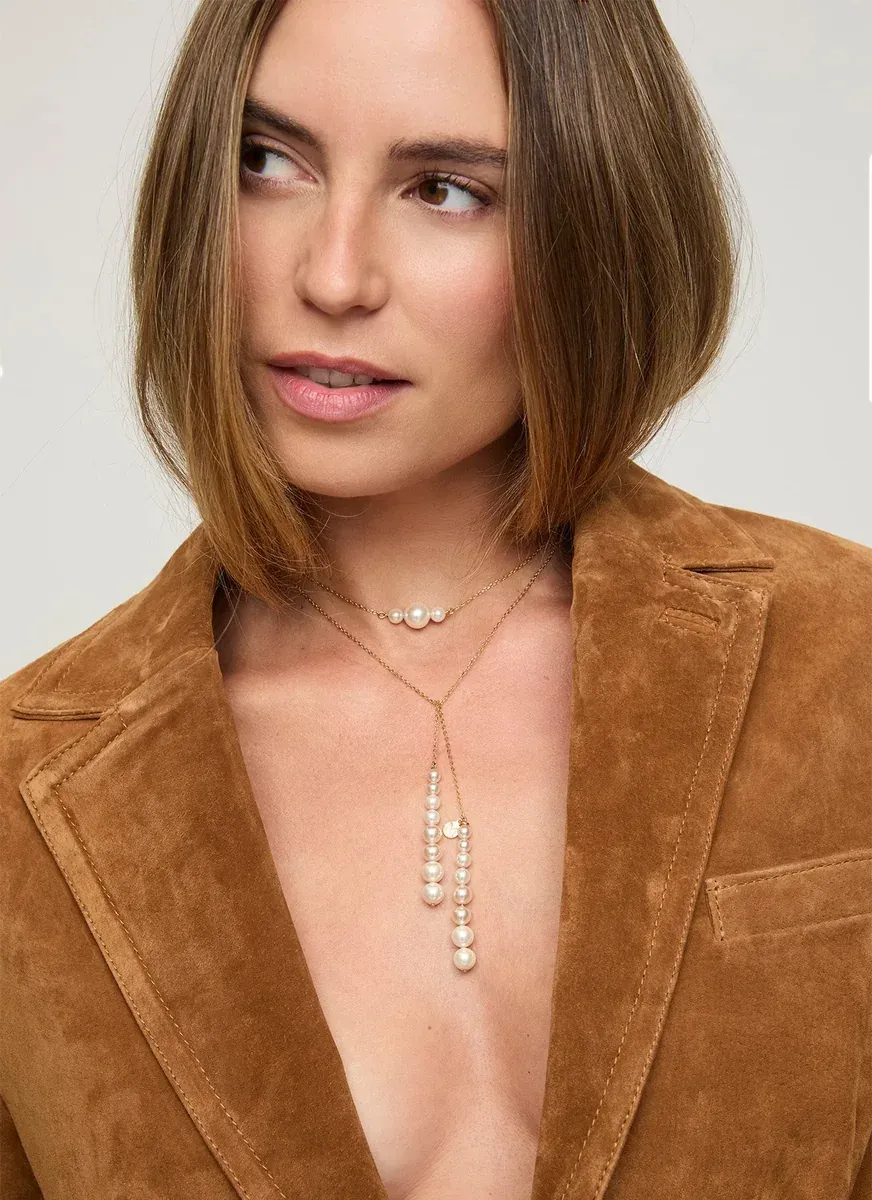 Primer plano del collar de perlas sobre el cuello de una modelo, con chaqueta de ante marrón, fondo neutro.