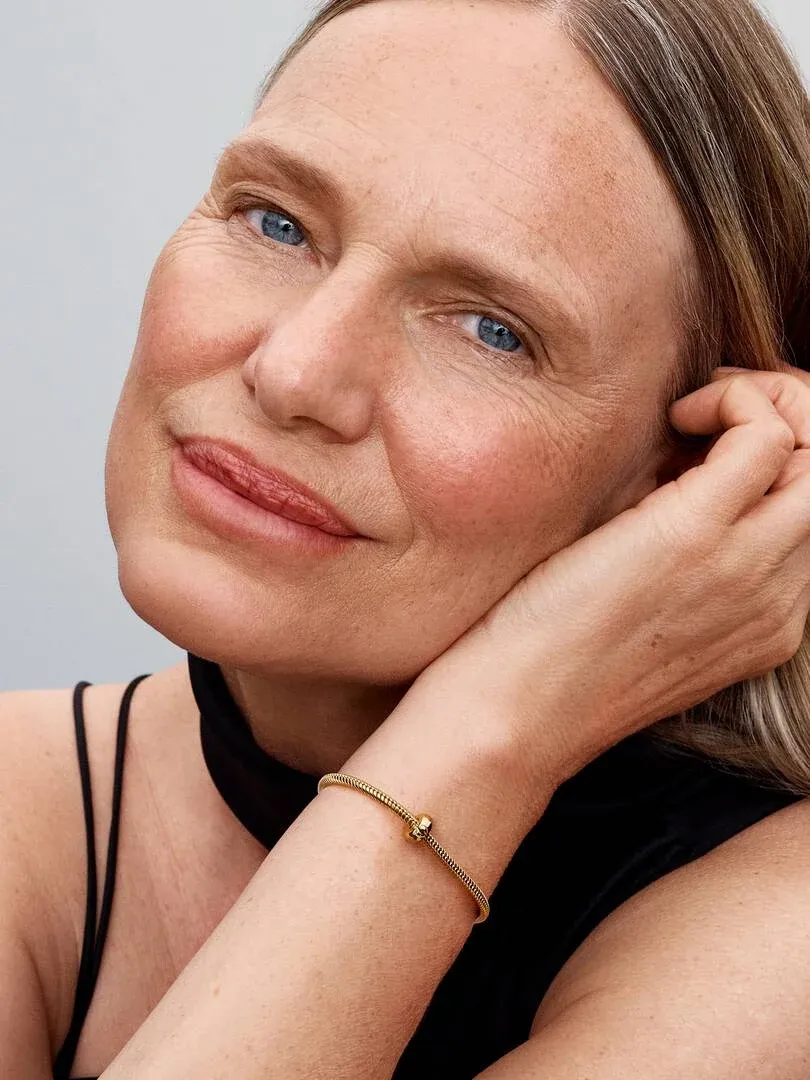 Close-up of gold bracelet on wrist, model resting face on hand, neutral background.