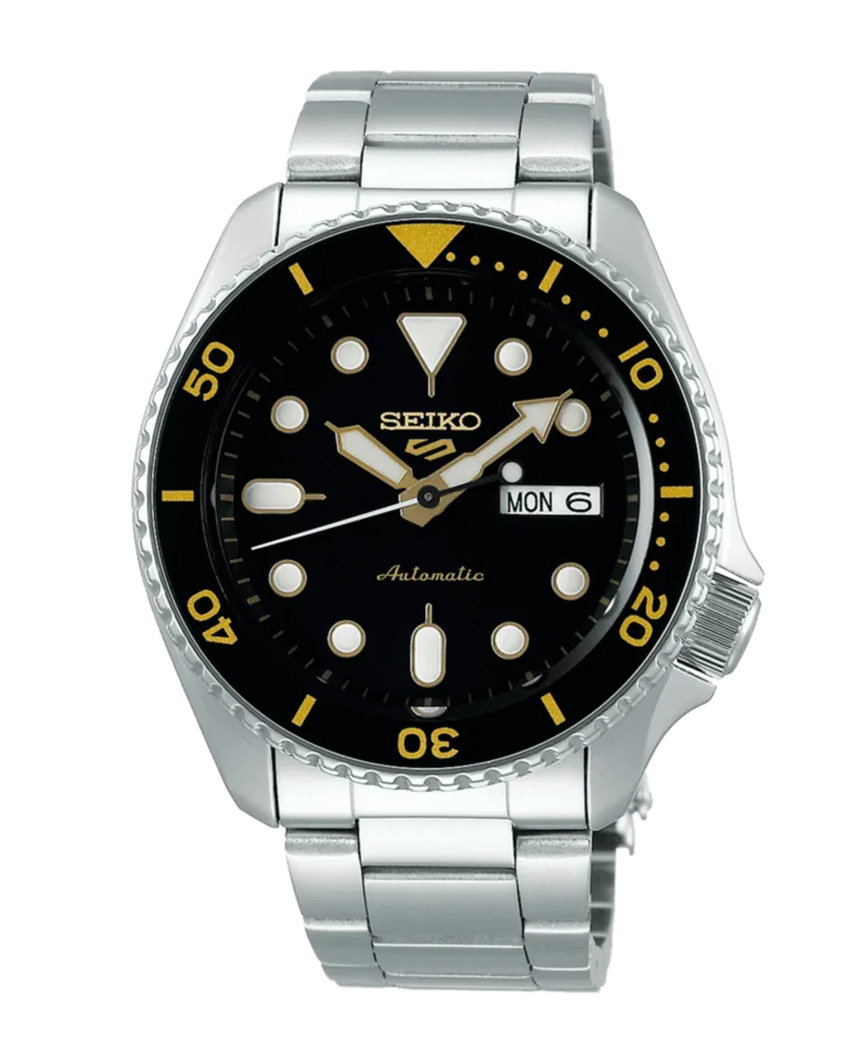 Vista frontal del reloj con esfera negra y detalles dorados, brazalete de acero inoxidable, fondo blanco.