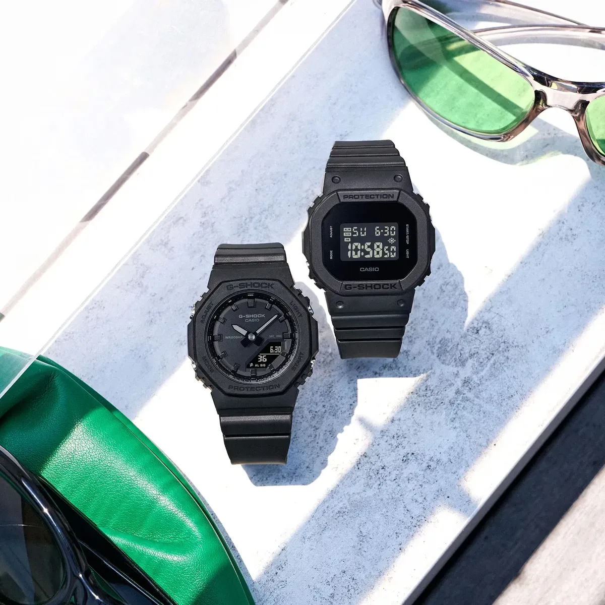 Vista cenital de dos relojes negros sobre superficie de mármol blanco, acompañados de gafas y accesorio verde.