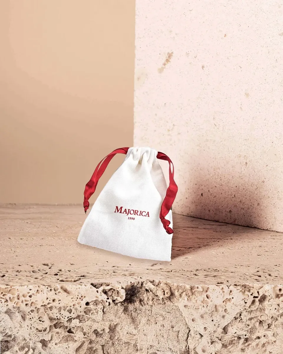 Bolsa de tela blanca con logo rojo de Majorica, situada sobre una superficie de piedra porosa, fondo neutro.