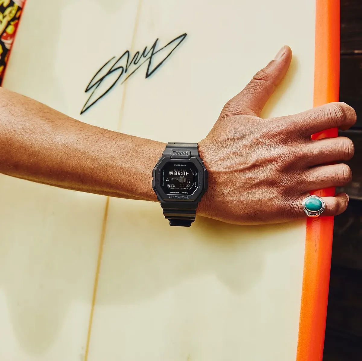 Reloj puesto en la muñeca de un modelo que sujeta una tabla de surf blanca con borde naranja.