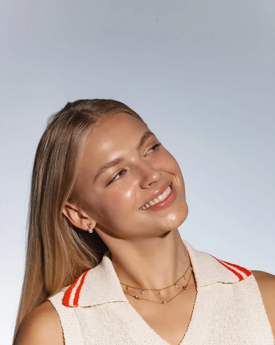 Primer plano de modelo sonriente con collar de perlas y detalles florales, toma lateral sobre fondo neutro.