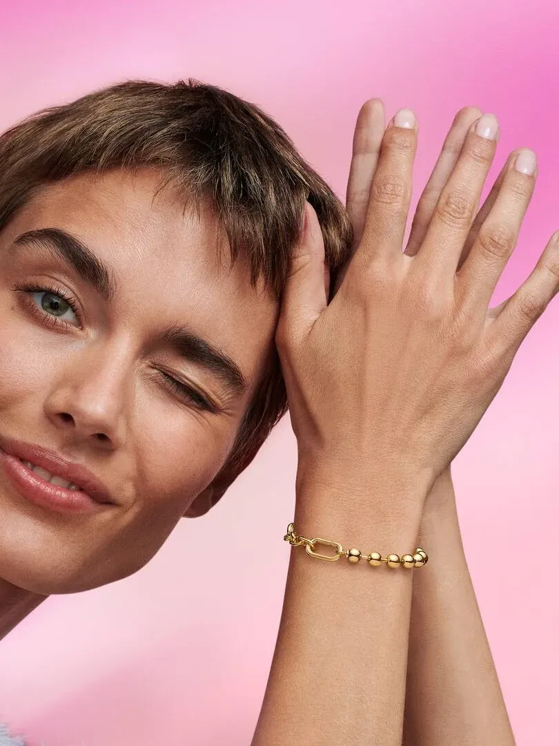 Nahaufnahme des goldenen Armbands am Handgelenk, Modell zwinkert vor rosa Hintergrund.