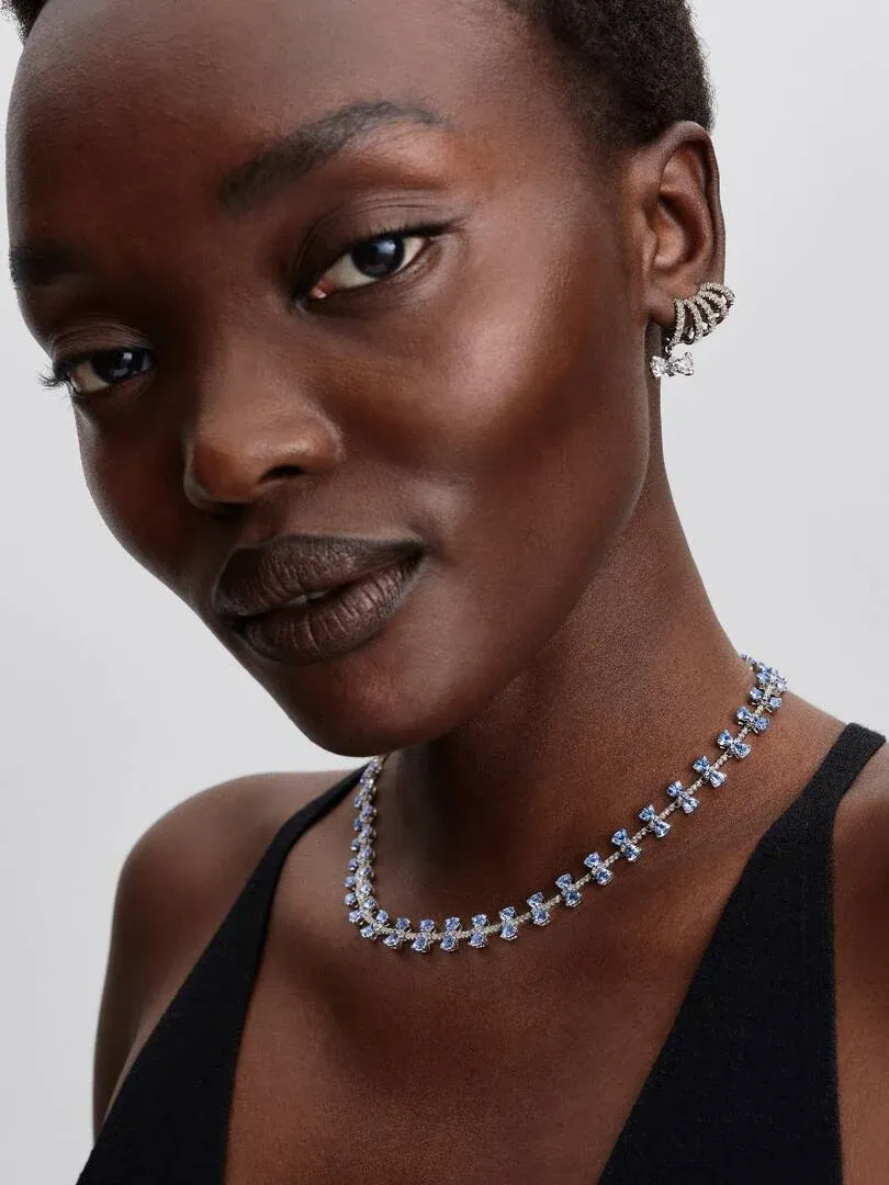 Close-up van model met de blauwe strikketting en bijpassende oorbellen tegen een licht neutrale achtergrond.