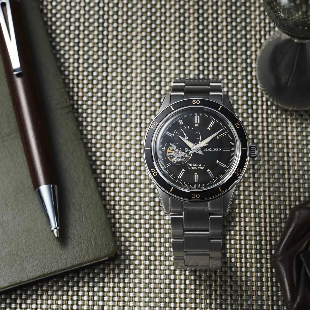 Vista cenital del reloj en composición con libreta de cuero y bolígrafo sobre superficie texturizada.
