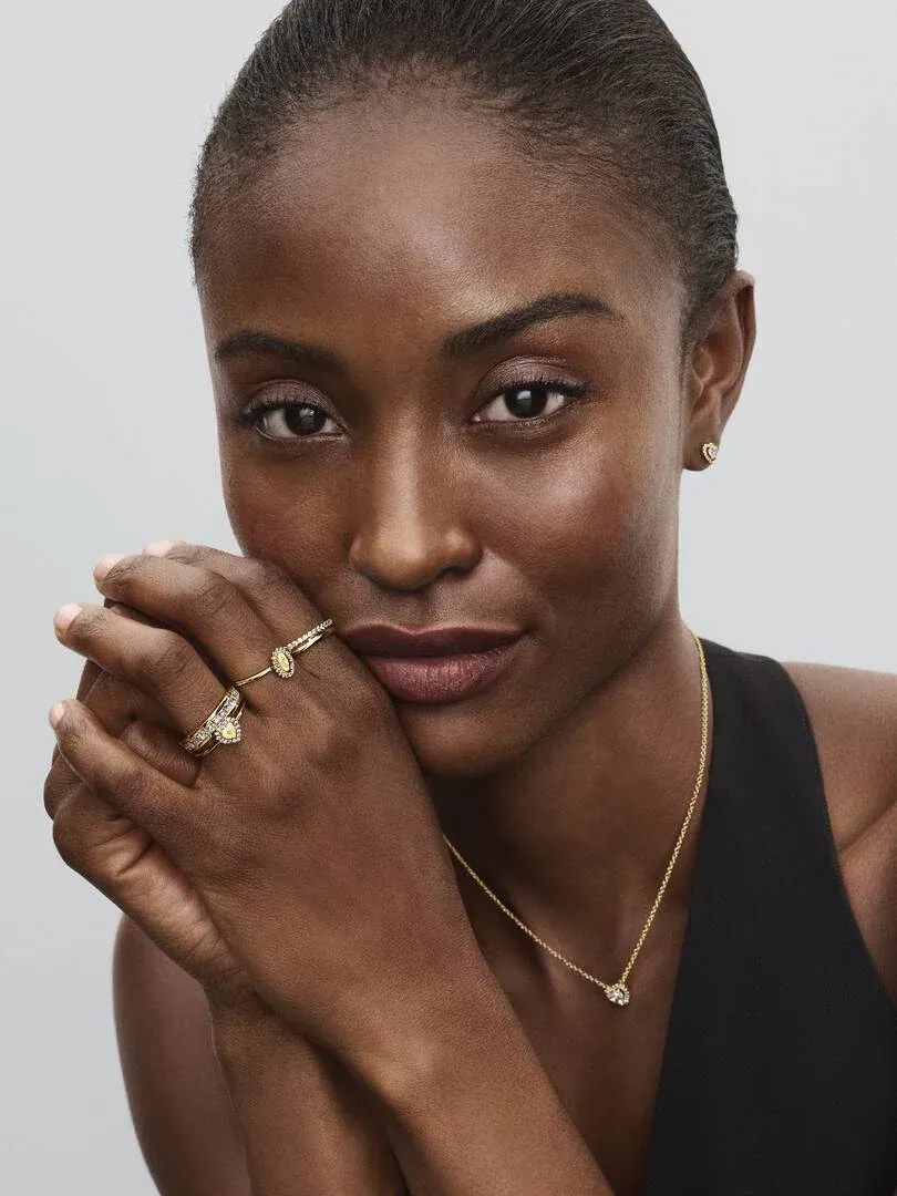 Nahaufnahme eines Models mit goldener Halskette, Ohrringen und Ring vor hellem, neutralem Hintergrund.