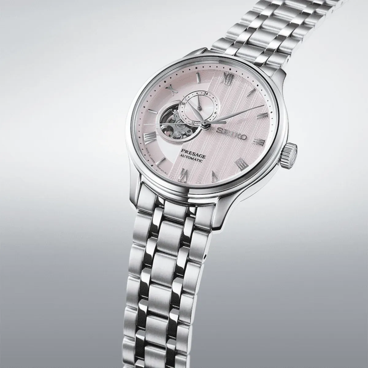 Three-quarter view of the watch on a neutral gradient background, showing the pink dial and steel bracelet.