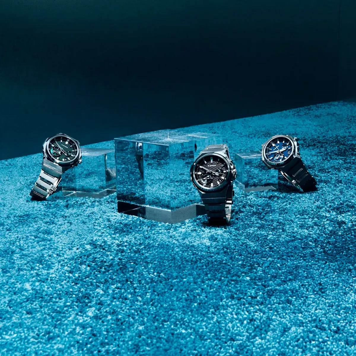Tres relojes de diferentes colores dispuestos sobre bloques de acrílico y una superficie azul texturizada.