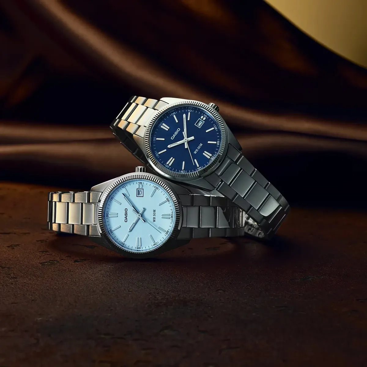 Dos relojes de acero con esferas en azul claro y azul oscuro, dispuestos en composición sobre fondo marrón texturizado.