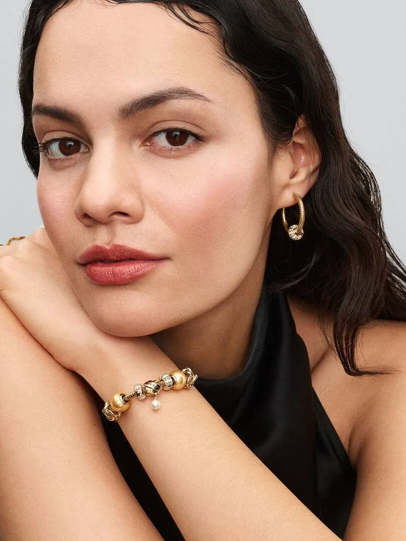 Nahaufnahme eines Models mit goldenen Creolen und Armband mit Charm, Frontansicht vor neutralem Hintergrund.