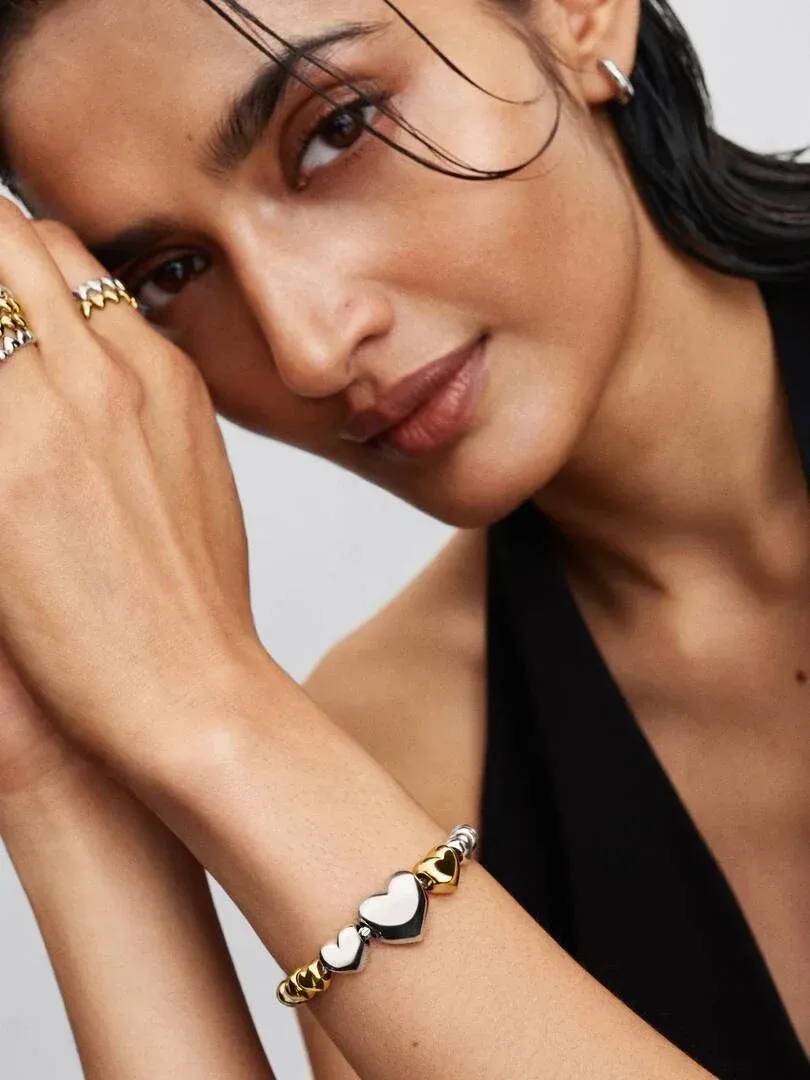 Nahaufnahme eines Handgelenks mit einem Charm-Armband, Modell posiert mit der Hand am Gesicht, neutraler Hintergrund.