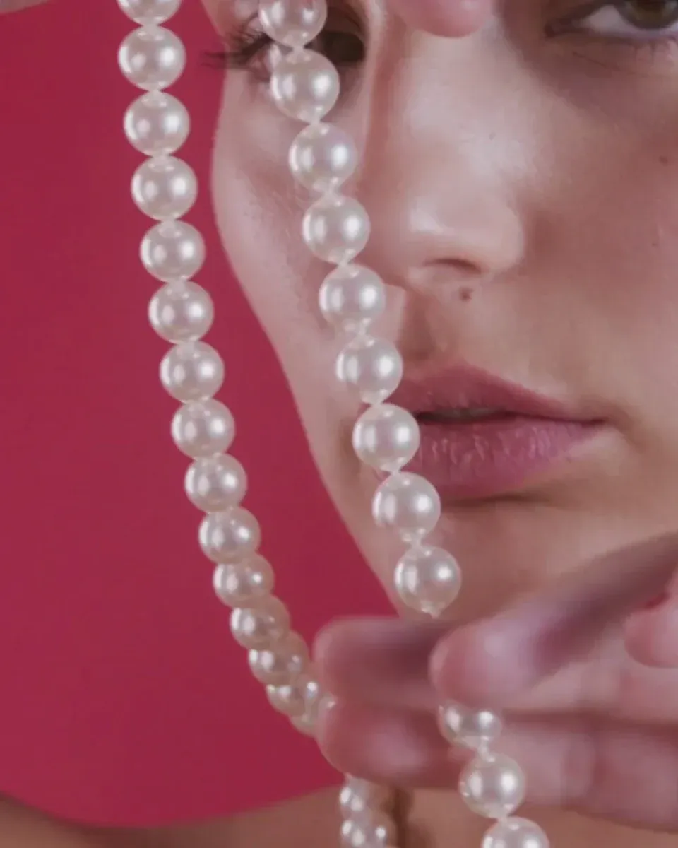 Close-up of white pearl necklace draped over a model's face, solid red background.