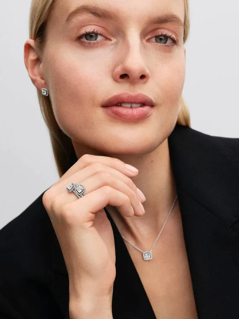 Nahaufnahme eines Models mit Halo-Halskette und Ohrringen, Hand am Gesicht vor neutralem grauem Hintergrund.