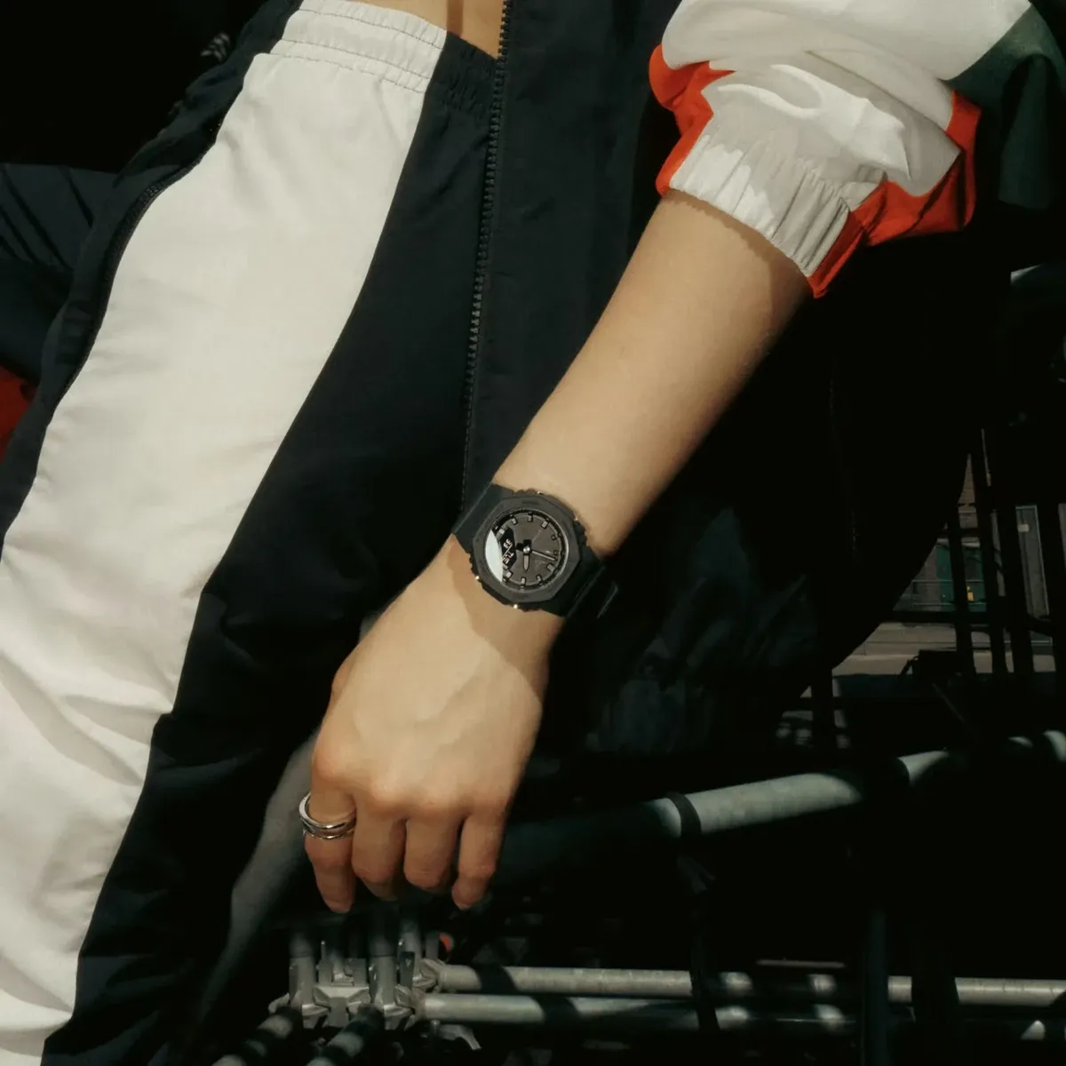 Reloj puesto en la muñeca de un modelo con chaqueta deportiva, fondo urbano con barandillas metálicas.