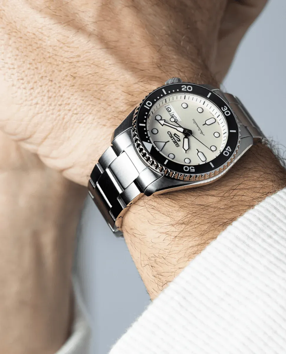 Montre portée au poignet, prise de vue en angle diagonal sur fond neutre flou, avec détail de manche blanche.