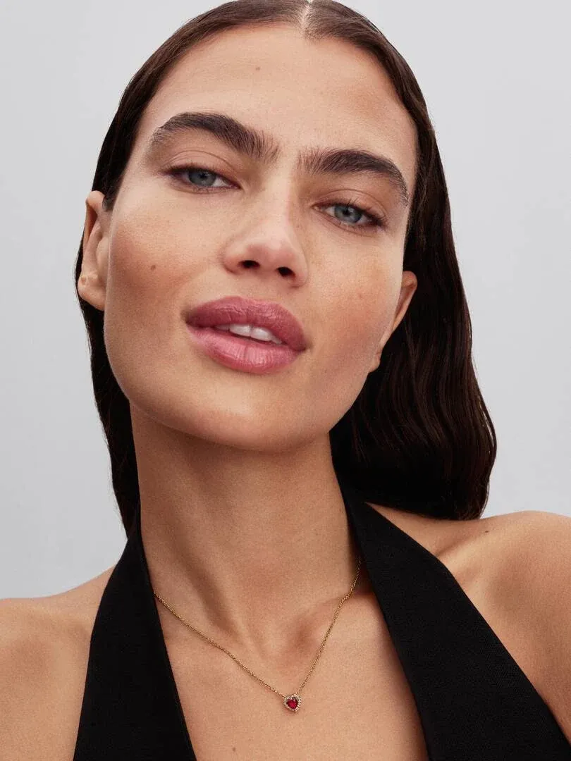 Primer plano del collar con colgante de corazón rojo sobre el cuello de una modelo, fondo neutro.