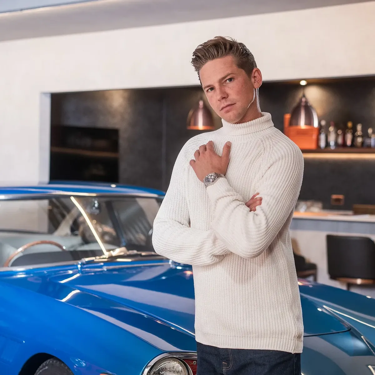 Hombre con jersey blanco y reloj en la muñeca, posando junto a un coche deportivo azul en un interior moderno.