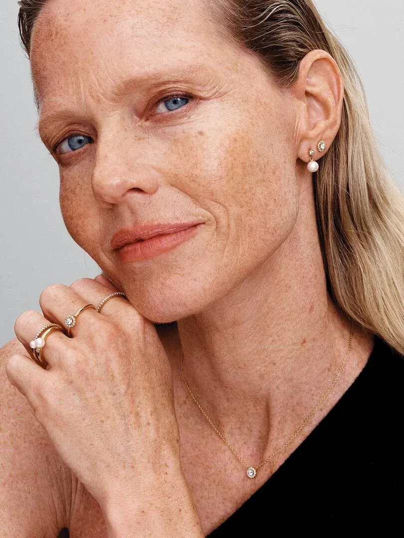 Nahaufnahme eines Models mit goldenen Ringen, Ohrringen und einer Halskette vor neutralem Hintergrund.