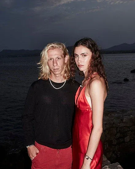 Dos modelos frente al mar al atardecer; primer plano del collar de perlas blancas sobre el cuello del modelo masculino.