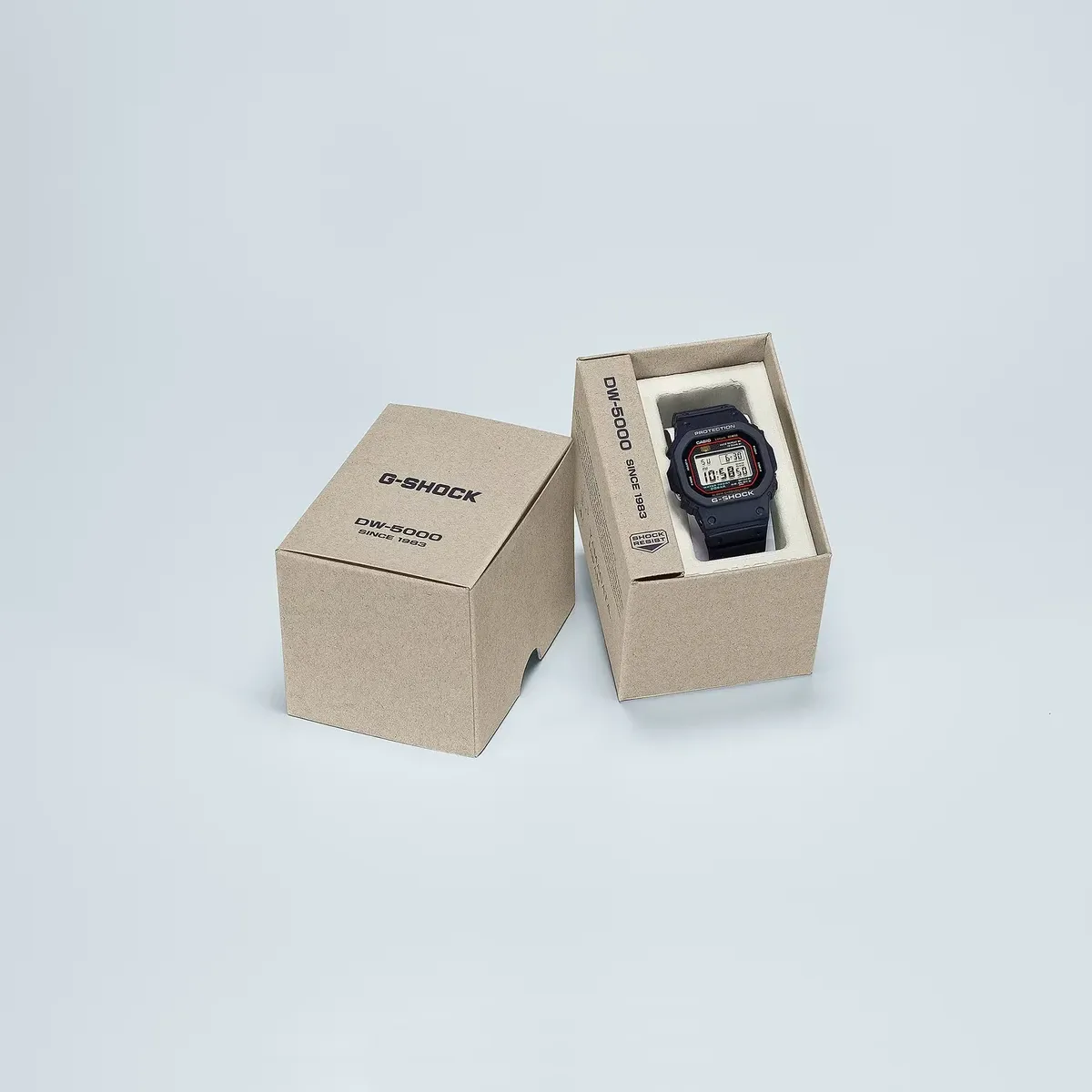 Reloj presentado en su caja de cartón kraft marrón con la tapa al lado, sobre fondo neutro gris claro.