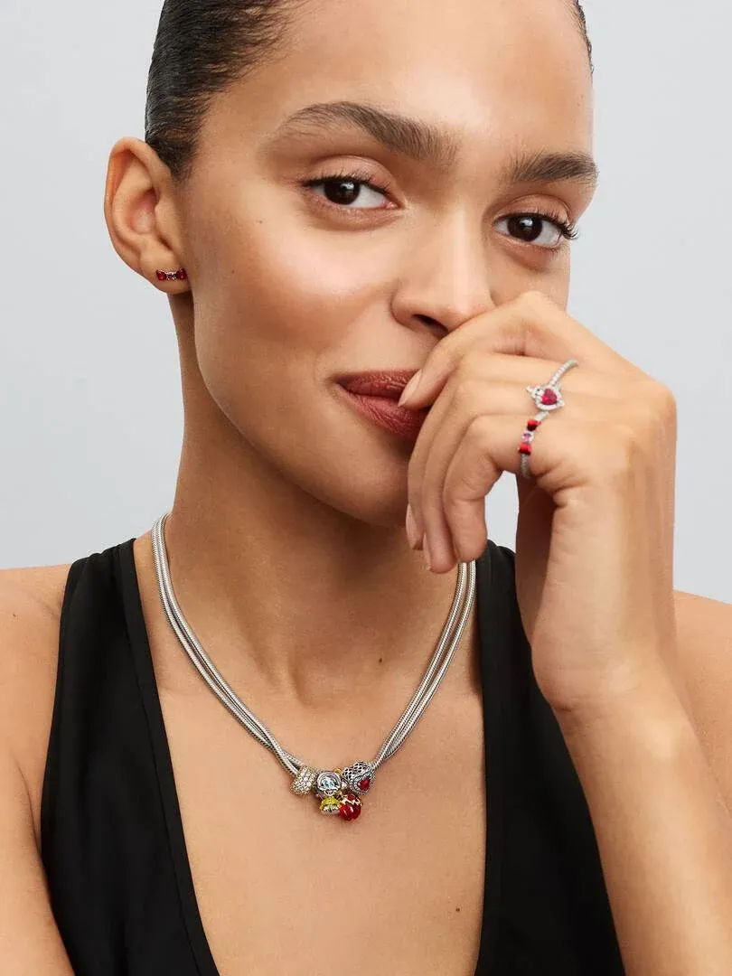 Model wearing silver necklace with colorful charm, red earrings, and matching ring against neutral grey background.