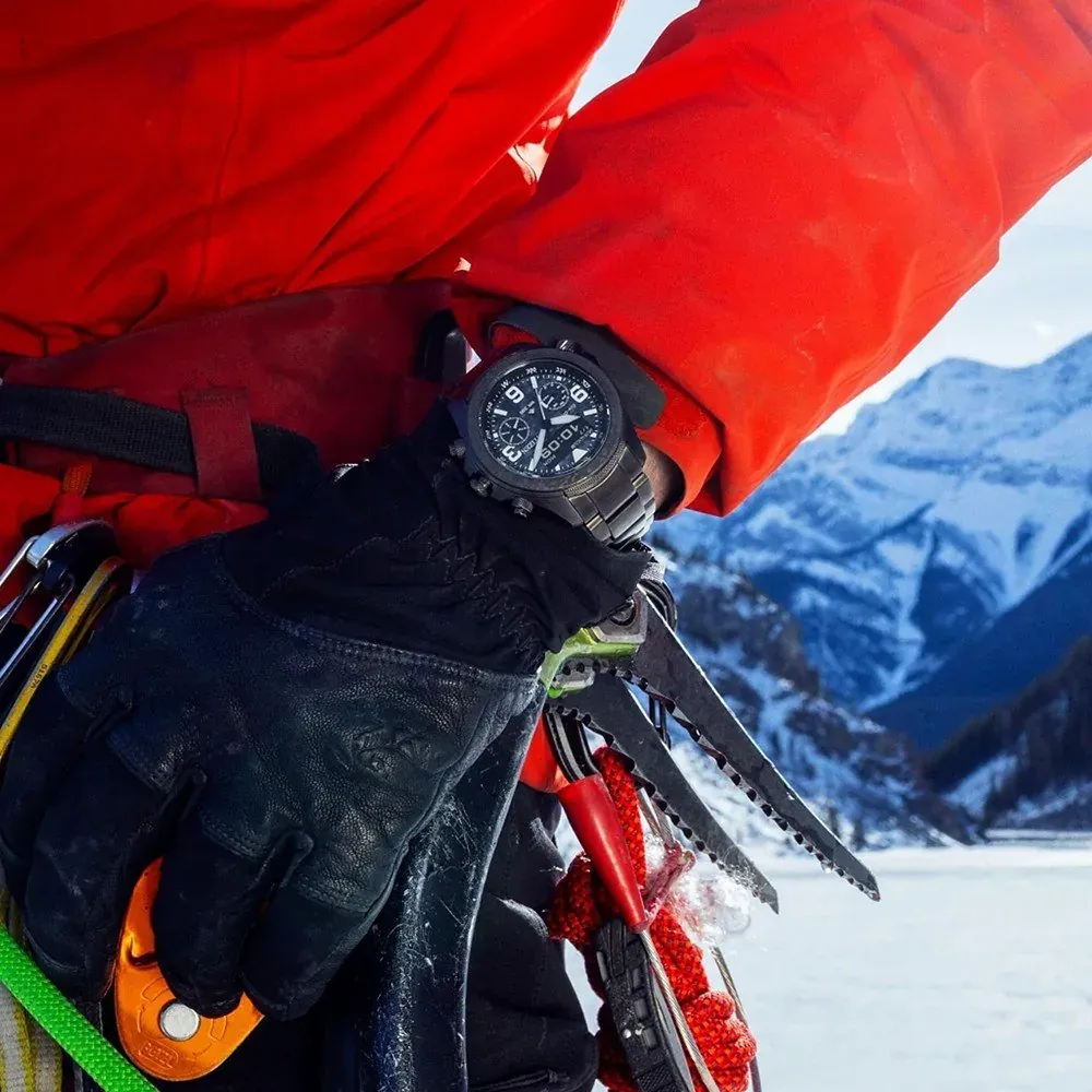 Reloj puesto en muñeca con guantes negros y chaqueta roja, fondo de montañas nevadas.