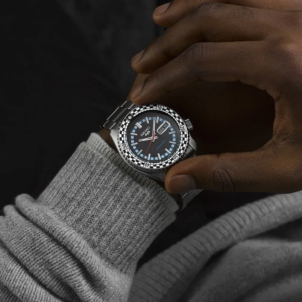 Watch worn on a male wrist with a grey sweater, hand touching the dial, dark background.