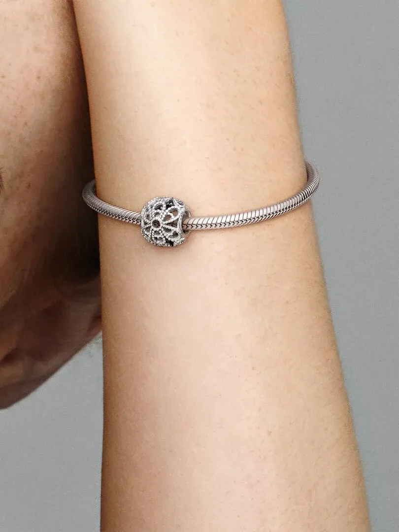 Close-up van een filigraan charm aan een zilveren armband, gedragen om de pols tegen een neutrale grijze achtergrond.