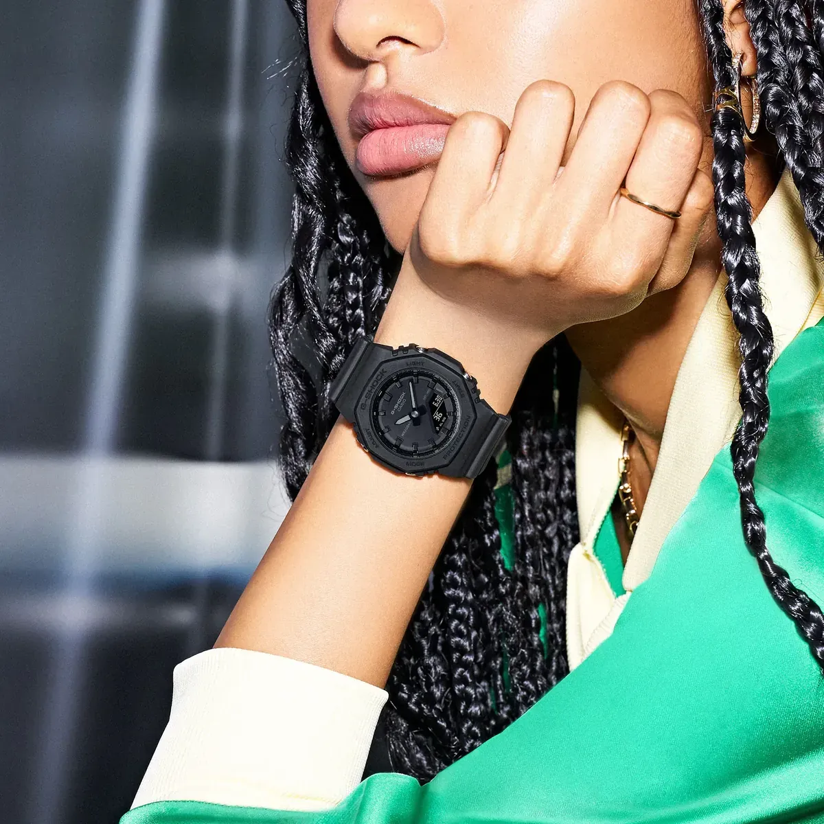 Primer plano del reloj negro en la muñeca de una modelo con trenzas, fondo desenfocado y ropa deportiva verde.