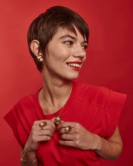 Model im roten T-Shirt mit Perlenhalskette, Ohrringen und Ring vor einfarbig rotem Hintergrund.