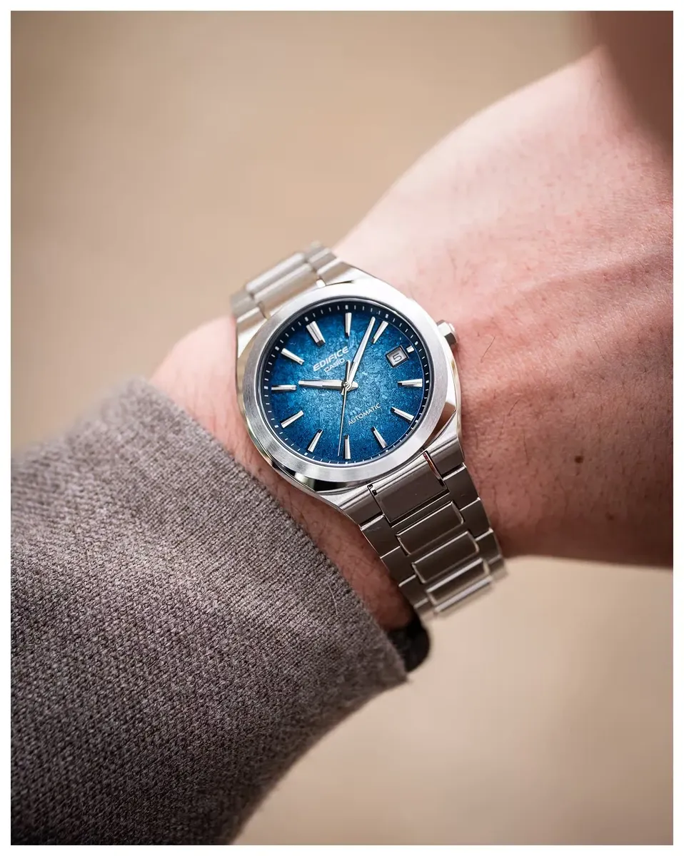 Reloj puesto en la muñeca con manga de jersey gris, toma en ángulo sobre fondo neutro desenfocado.