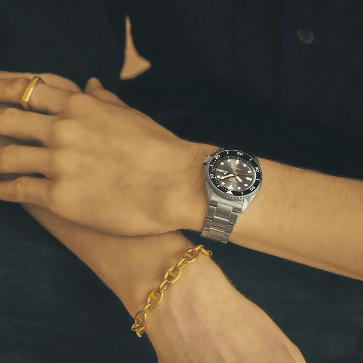 Watch on a man's wrist next to a gold link bracelet and a ring, blurred dark background.