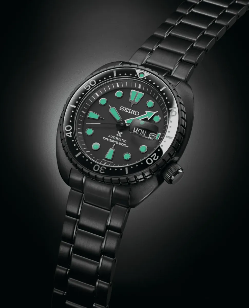 Reloj negro en ángulo cenital inclinado sobre fondo degradado oscuro, vista completa de la esfera y el brazalete.