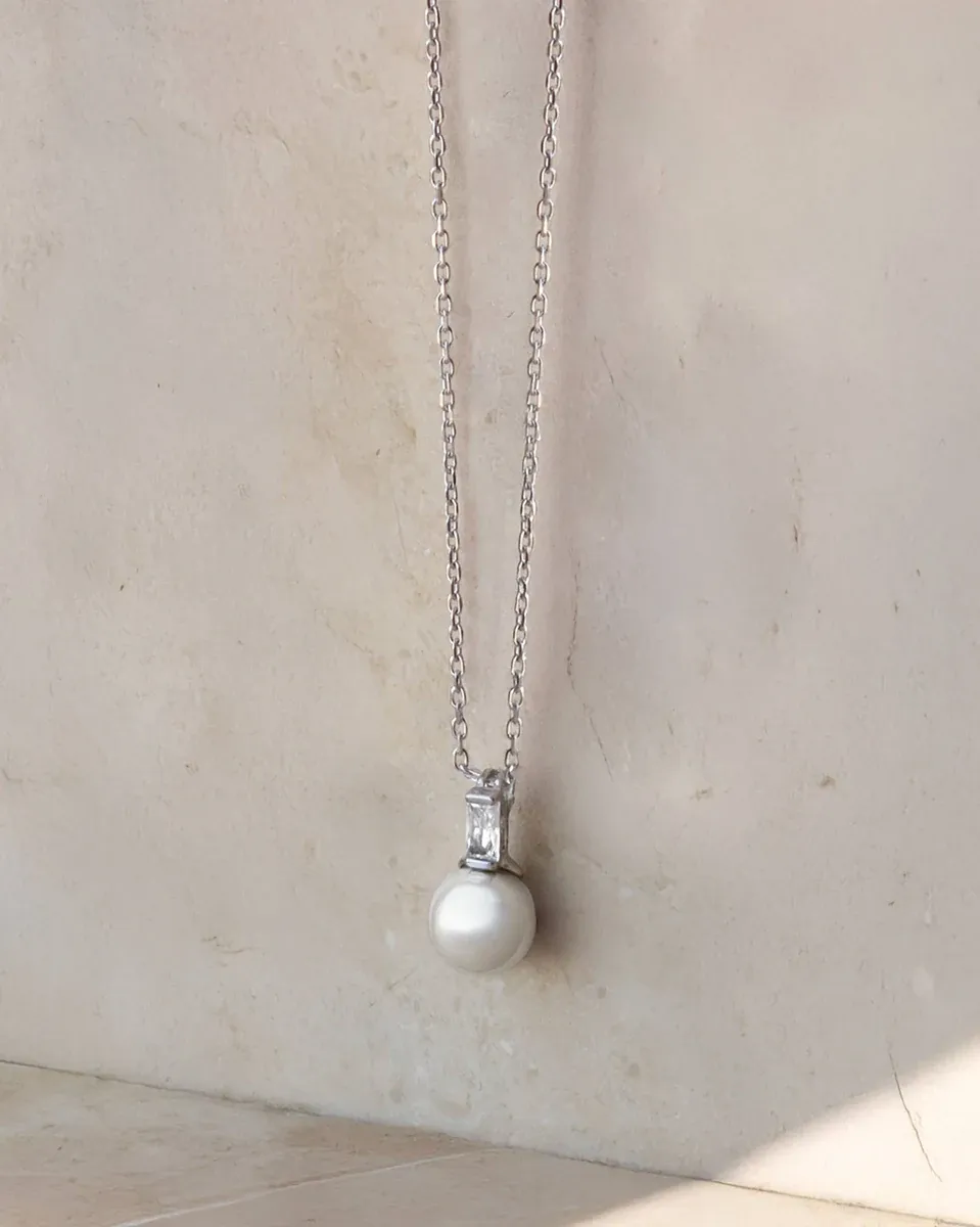 Colgante de perla blanca con cadena de plata, vista frontal sobre fondo neutro texturizado.