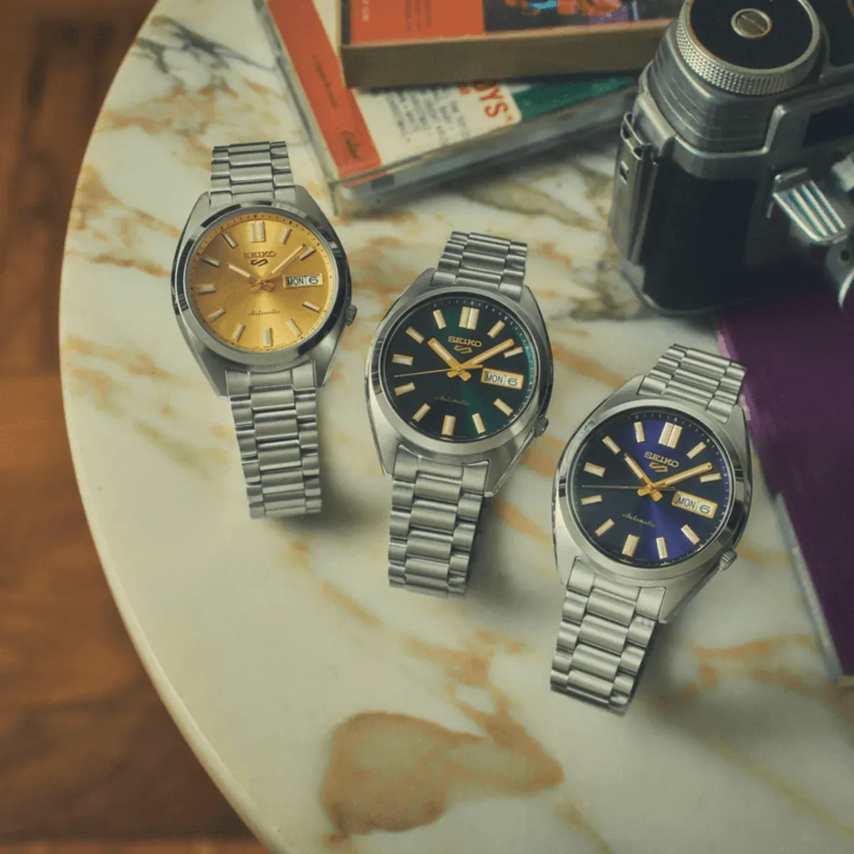 Trois montres avec cadrans de différentes couleurs sur table en marbre, à côté d'un appareil photo et de livres, vue de dessus.