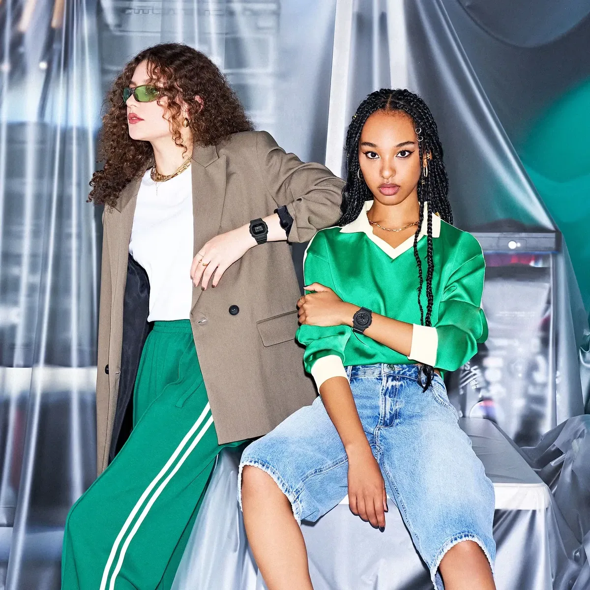 Dos modelos posando con relojes negros, vestidas con ropa casual y deportiva, fondo de cortinas plásticas blancas.