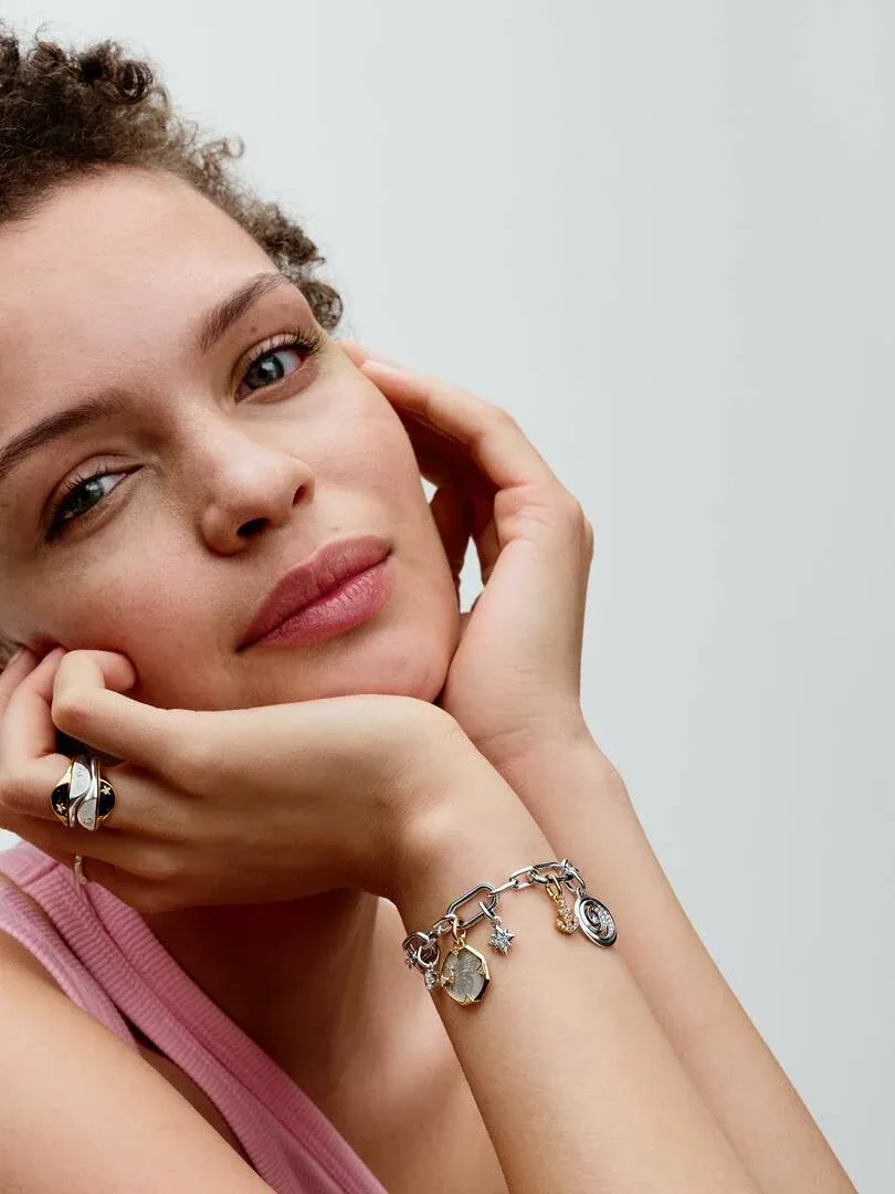 Close-up of model wearing charm bracelet and ring, front view with hands framing face, neutral background.