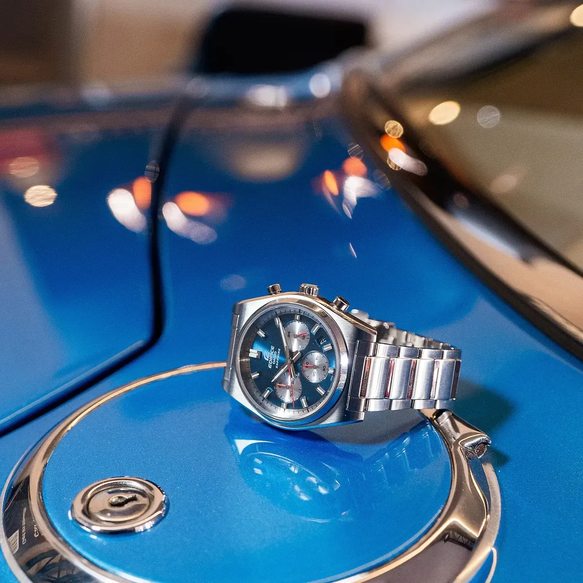 Reloj apoyado sobre el capó azul brillante de un coche clásico, toma en ángulo con reflejos metálicos.