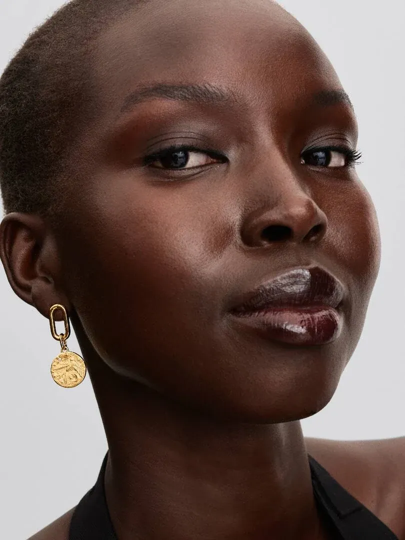 Zijaanzicht van gouden medaillon oorbel gedragen door een model tegen een lichte neutrale achtergrond.