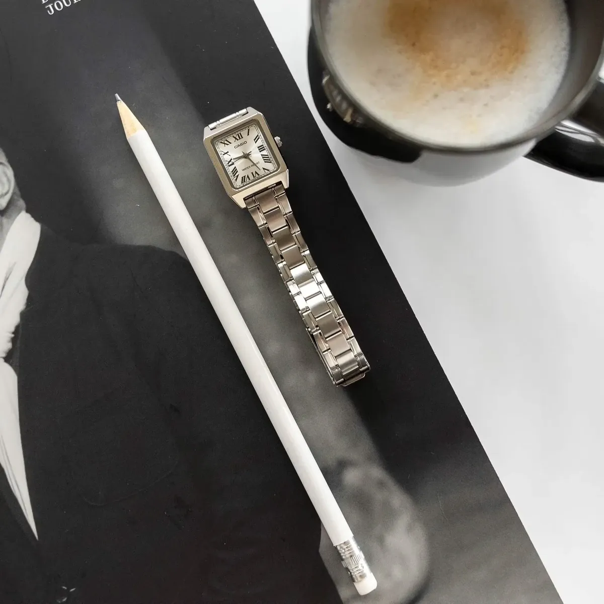 Composición cenital de reloj cuadrado junto a un lápiz blanco y una taza de café sobre una revista en blanco y negro.