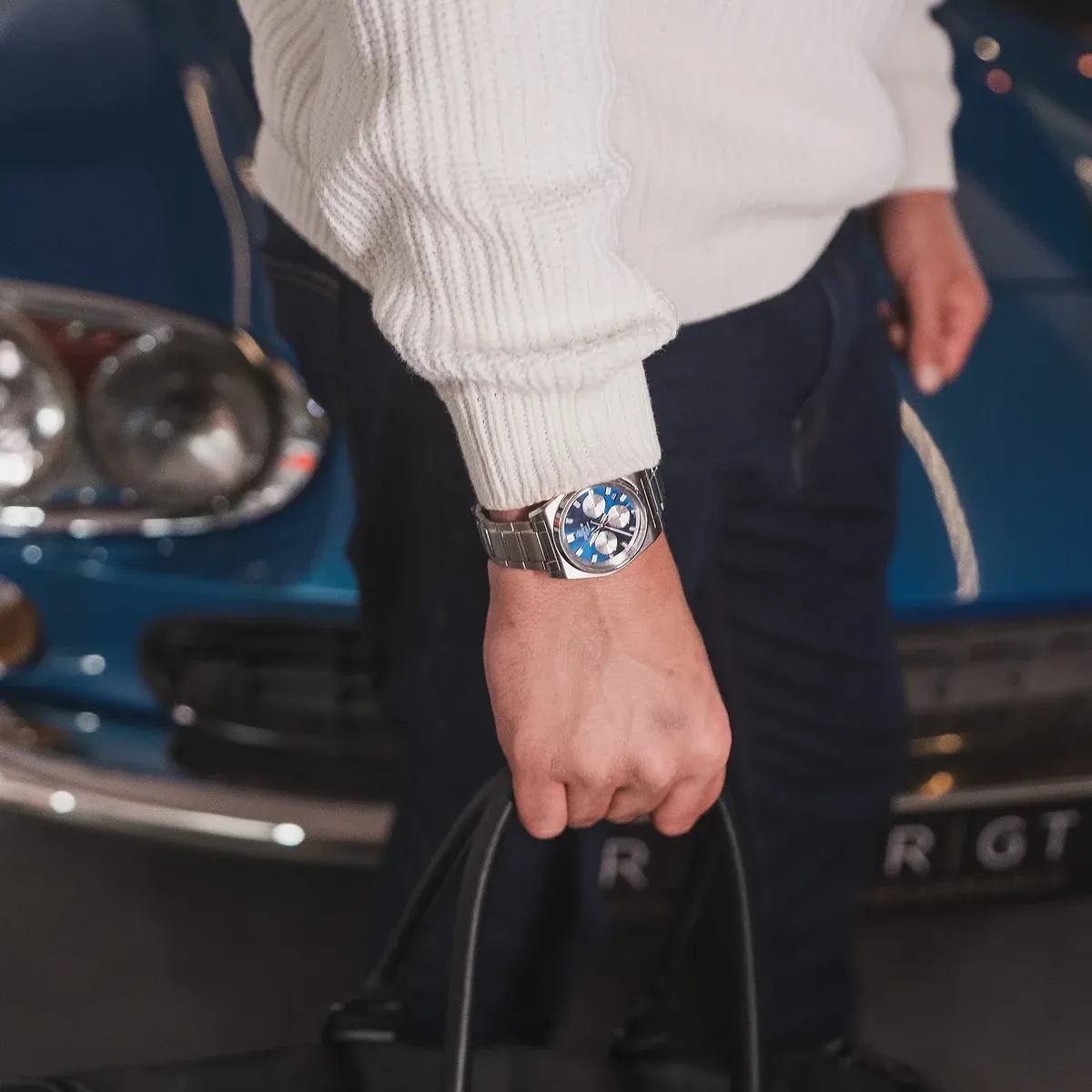 Primer plano del reloj puesto en la muñeca, mano sosteniendo un bolso negro, fondo de coche azul.