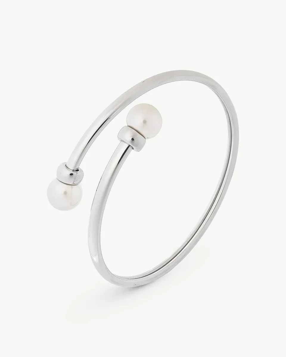 Brazalete rígido de acero con dos perlas blancas en los extremos, vista en ángulo sobre fondo blanco.