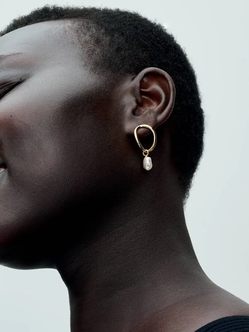 Gros plan latéral d'une boucle d'oreille perle baroque portée à l'oreille, fond neutre clair.