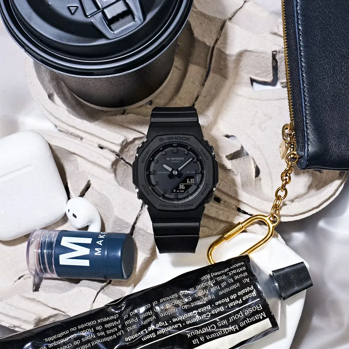 Composición cenital del reloj negro rodeado de accesorios cotidianos sobre fondo claro texturizado.
