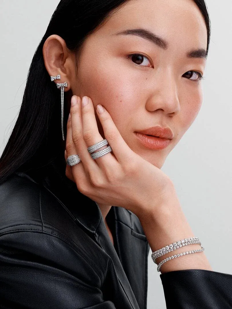 Nahaufnahme eines Models mit Hängeohrringen und Tennisarmband, neutraler Hintergrund.